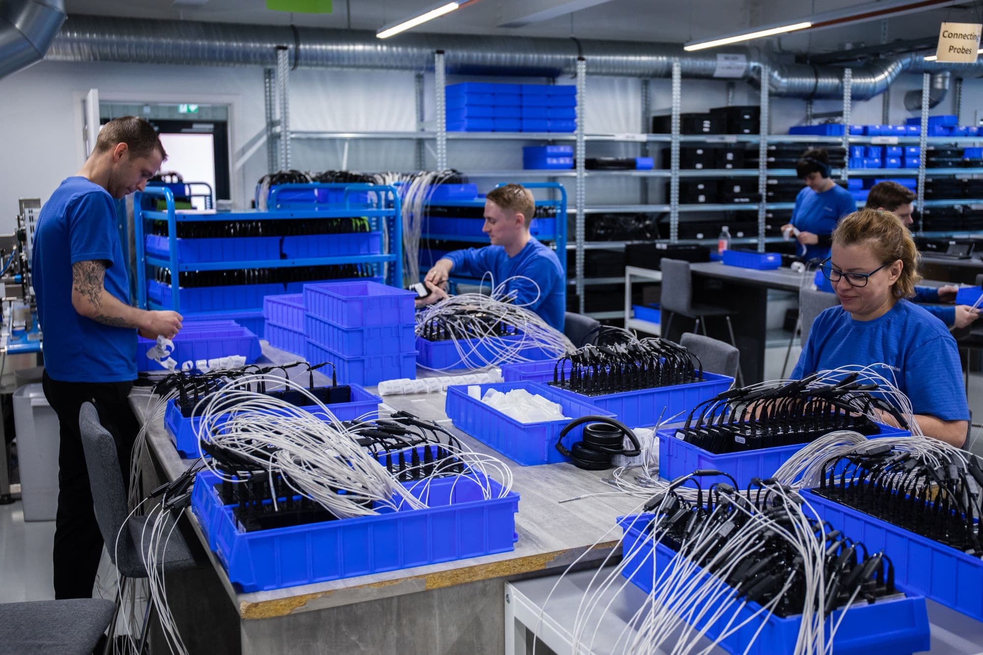Controlant staff in blue shirts at workbenches in Reykjavik, connecting probe harnesses to crates of Saga devices, with racks of more behind them