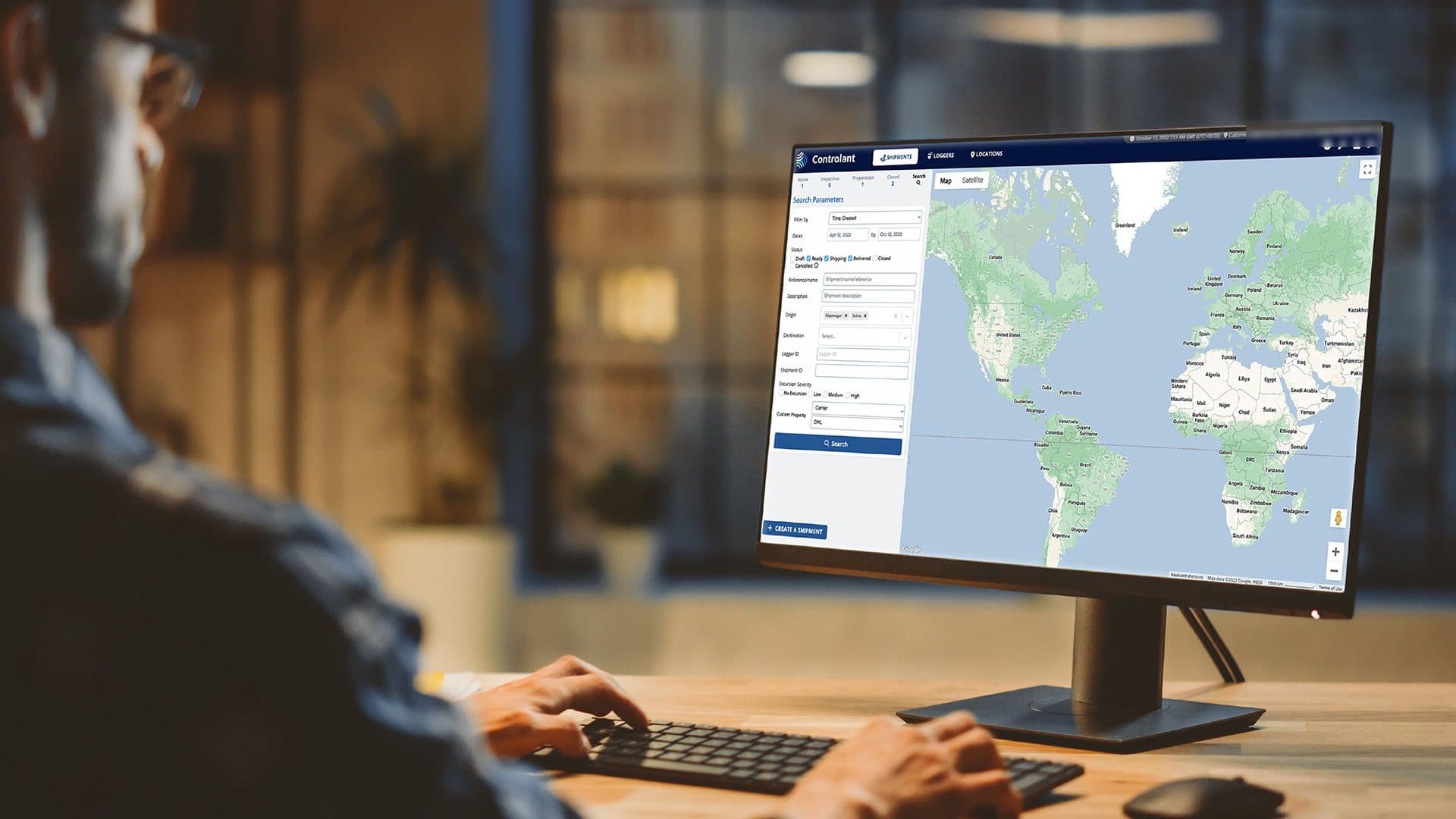 A man seen from behind at a desktop monitor in an evening office, the screen showing the platform's shipment search and a world map