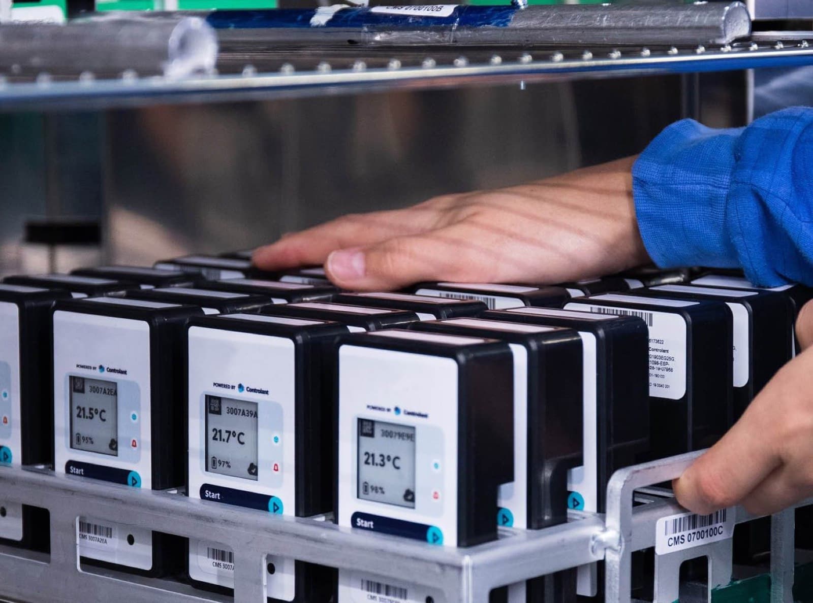 Hands at a metal rack of Saga devices, their screens reading 21.3 to 21.7 ยฐC